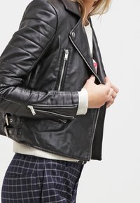 Černá kožená motorkářská bunda s prošitými detaily na ramenech, asymetrickým zapínáním na zip a dvěma kapsami na zip. Měkká textura a přiléhavý střih.