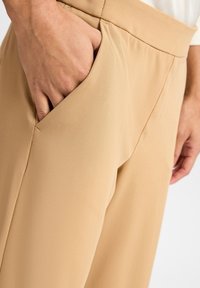 Pantalon beige à texture lisse, doté d'une poche latérale et d'une coupe ample, présenté de la taille jusqu'en bas avec une main reposant dans la poche.