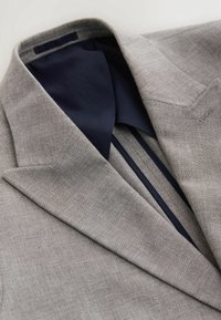 Veste de costume gris clair avec un col cranté et une doublure intérieure bleu marine foncé visible le long du col et de la couture intérieure.