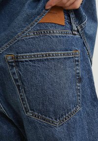 Vaqueros de mezclilla azul con un acabado texturizado, presentan un bolsillo trasero y un parche de cuero marrón con texto en relieve. Se puede ver el pespunte de contraste.