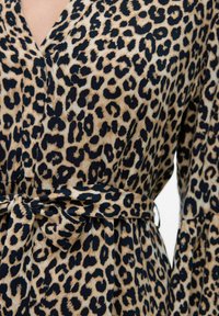 Robe imprimée léopard en beige et noir, dotée d'un décolleté en V, de manches amples et d'une ceinture nouée à la taille. Tissu lisse avec une texture douce.