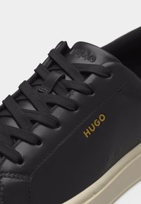 Κοντινή λήψη μαύρου δερμάτινου αθλητικού παπουτσιού με μαύρα κορδόνια, μπεζ σόλα και χρυσό λογότυπο "HUGO" στο πλάι κοντά στην περιοχή των κορδονιών.