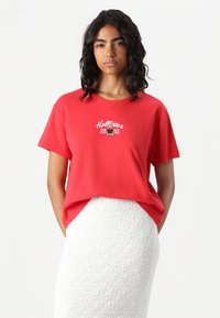 Red cotton t-shirt featuring front graphic text "Hollister 2002" with a relaxed fit and short sleeves, paired with a textured white skirt.
