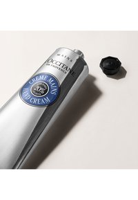 Metaliczna srebrna tubka kremu do rąk z niebieską etykietą z napisem "Crème Mains" oraz czarną zakrętką obok. Gładka tekstura, owalny kształt.