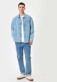 Giacca di jeans azzurro chiaro con chiusura a bottoni, tasca sul petto e vestibilità ampia, abbinata a jeans azzurri chiari e scarpe da ginnastica bianche.