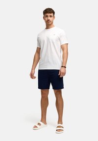 Camiseta de algodón blanca con bolsillo, pantalones cortos texturizados en azul marino y sandalias blancas. El modelo está de pie de frente contra un fondo neutro.