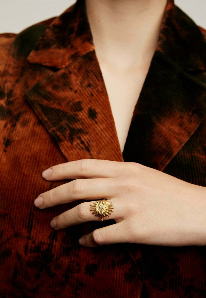 Anillo de oro con un diseño de destello solar, que presenta un centro ovalado y garras radiantes. Lleva puesto en una mano que reposa sobre una tela de sarga marrón texturada.