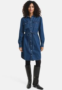 Denim shirt dress with a button front, chest pockets, and a tied waist. Features long sleeves and a knee-length design. Worn with tall black boots.