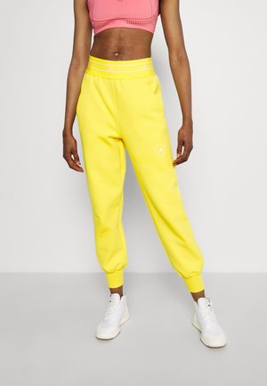 Kvinde iført lyse gule joggerbukser, lyserød sports-bh og hvide sneakers, stående mod en ensfarvet lys baggrund.