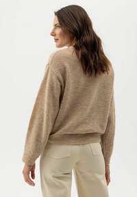 Homokszínű kötött pulóver, laza fazonnal, texturált felülettel, esővállakkal és bordázott aljjal. Világos színű nadrággal párosítva.