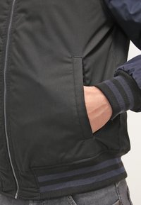 Personne portant une veste noire avec un poignet rayé, main insérée dans une poche avant, et une fermeture éclair argentée visible sur le côté gauche.