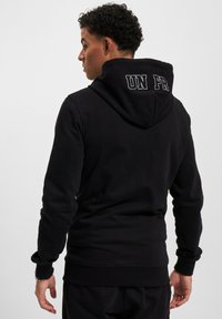 Maglia con cappuccio nera, con maniche e orlo a coste, che presenta "UN FR" ricamato in bianco sul retro. Tessuto morbido e texture.