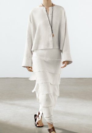 Lightweight, long-sleeve, cream linen top with a buttoned neck and loose fit, paired with a tiered, frayed white skirt and brown sandals.