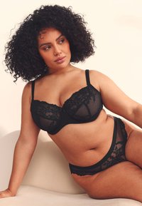 Femme aux cheveux bouclés portant un soutien-gorge en dentelle noire et des sous-vêtements assortis, assise et regardant vers sa gauche sur un fond uni.