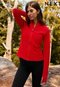 Red zip-up athletic jacket with long sleeves and a fitted design. Made of smooth fabric, featuring a stand-up collar and thumbholes.