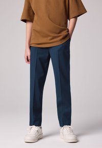 Ruskea oversize T-paita, jossa on lyhyet hihat, yhdistetty tummansinisiin räätälöityihin housuihin ja valkoisiin tennareihin. Sileä kangas ja rento istuvuus.