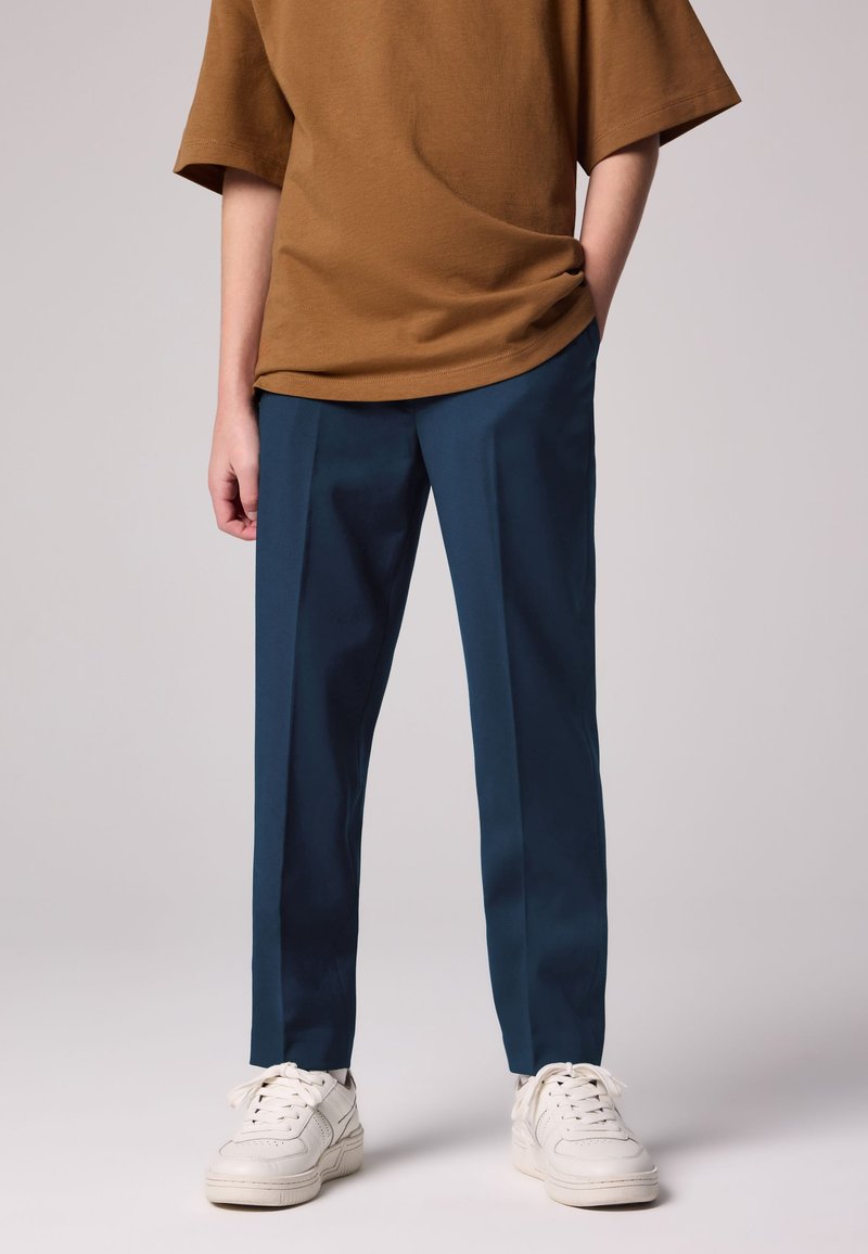 Ruskea oversize T-paita, jossa on lyhyet hihat, yhdistetty tummansinisiin räätälöityihin housuihin ja valkoisiin tennareihin. Sileä kangas ja rento istuvuus.