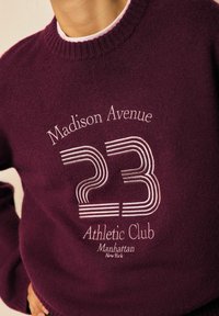 Bordo neule, jossa on valkoista brodeerattua tekstiä "Madison Avenue 23 Athletic Club Manhattan New York" rinnassa, päällä oleva henkilö.