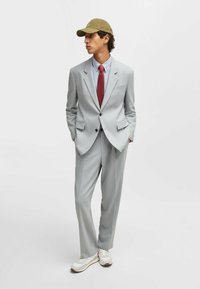 Light gray suit with slim fit, notched lapels, single-button closure; paired with a blue shirt, red tie, and olive cap, white sneakers.
