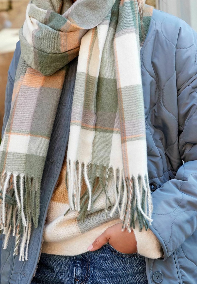 Personne portant une veste matelassée bleue, un pull beige, un jean bleu et une grande écharpe à carreaux avec des franges dans des tons verts, blancs et orange.