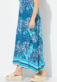 Longue robe bleue ornée de motifs variés dans des nuances de bleu et de blanc, avec un ourlet rayé, portée avec des sandales compensées beiges.