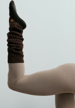 Une personne assise plie une jambe vers le haut, portant des collants beige, un chauffe-jambes côtelé foncé et une chaussure noire texturée sur un fond pâle.