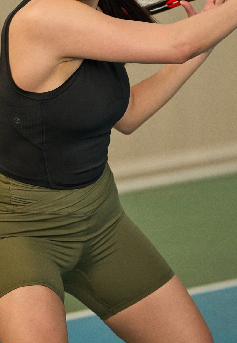 Personne portant un haut sans manches noir et un short vert olive, tenant une raquette de badminton rouge et noire dans un environnement sportif intérieur.