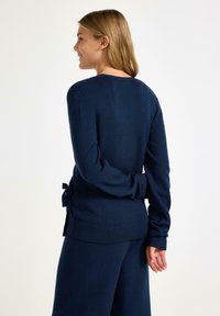 Marineblå strikket pullover med lange ærmer, ribbet kant og bindebånd på ryggen. Blød tekstur, tætsiddende design.