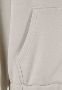 Light beige sweatshirt fabric with a front pocket. Features visible stitching and a gathered waistband at the bottom. Soft texture.