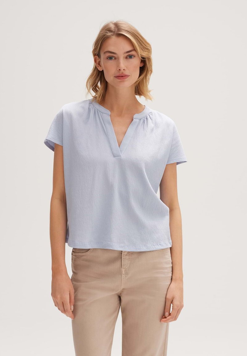 Blusa azzurro chiaro a maniche corte con scollatura a V, caratterizzata da sottili pieghe sul colletto e una vestibilità morbida. Il materiale sembra morbido e testurizzato.