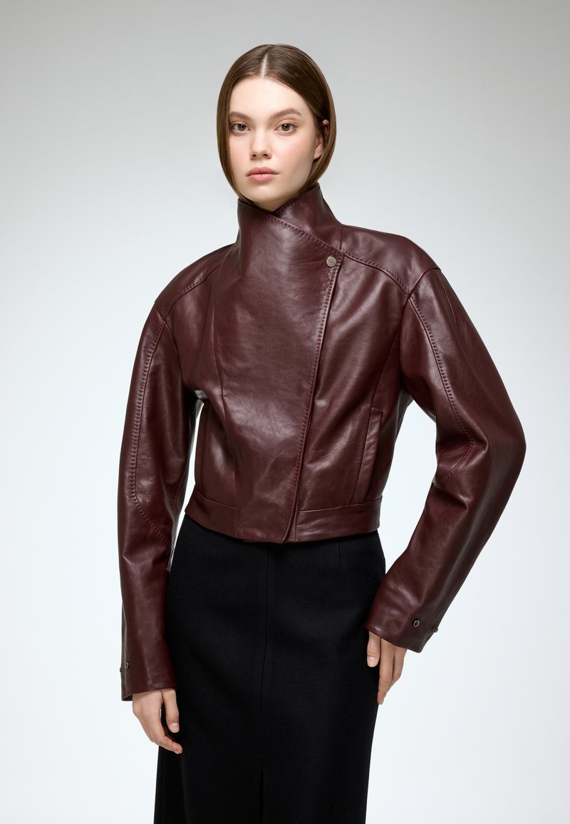 Burgundy leather jacket with a high collar, asymmetrical zip, and long sleeves, paired with a black skirt. Smooth texture, tailored fit.
