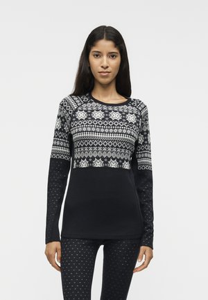 Γυναίκα με μακριά σκοτεινά μαλλιά, που φοράει μακρυμάνικη μαύρη και άσπρη μπλούζα με σχέδια και μαύρο κολάν με μικρά λευκά σχέδια, στέκεται μπροστά σε λευκό φόντο.