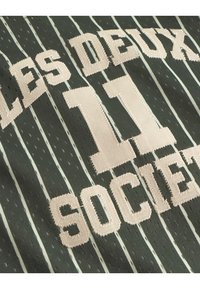 Sötétzöld szövet, függőleges fehér csíkokkal, bézs hímzett szöveggel: "LES DEUX" és "11 SOCIETÉ" egy merész, sportos betűtípussal.