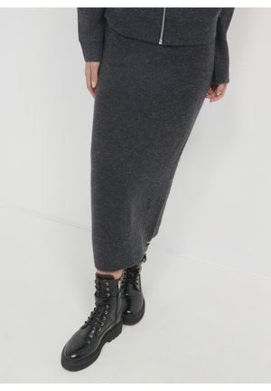 Personne portant une jupe midi en tricot gris foncé et un haut assorti, associé à des bottines noires à lacets brillantes sur fond blanc.