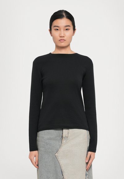 Woman wearing a black long-sleeve shirt and a two-tone denim skirt, standing against a plain white background, facing forward.