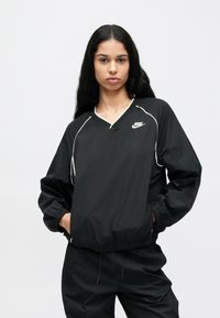 Fekete Nike pulóver kabát V-nyakú, fehér szegélydíszítéssel, elasztikus mandzsettákkal és laza illeszkedéssel. A modell egyszínű háttér előtt áll.