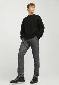 Zwarte gestructureerde trui, grijze broek en zwarte schoenen. De trui heeft een ronde hals, een ruime pasvorm en een gebreid patroon. De broek is op maat gemaakt.