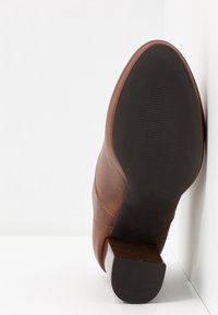 Bottine en cuir marron avec un talon bloc, semelle tournée vers l'avant, appuyée contre un angle entre un mur blanc et le sol.