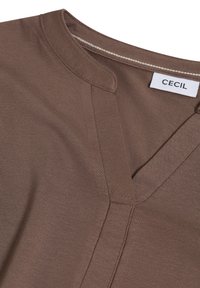 Haut en coton marron avec un décolleté en V, une texture subtile et des détails cousus ; l'étiquette indique "CECI".