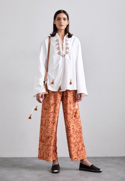 Blouse blanche avec des accents en franges et broderies marron. Pantalons amples orange avec un motif floral. Des ballerines tissées noires complètent la tenue.