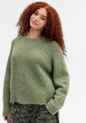 Junge Frau mit lockigem rotem Haar trägt einen flauschigen grünen Pullover und Camouflage-Hose, die Hand in der Tasche, lächelt sanft vor einfachem Hintergrund.
