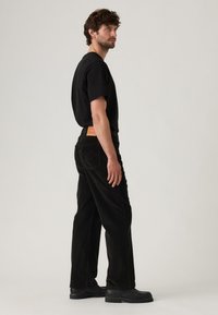 Man die zijwaarts staat, gekleed in een zwart t-shirt, zwarte corduroybroek met een leren patch en zwarte schoenen tegen een effen achtergrond.