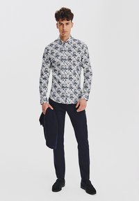 Biała koszula z kołnierzykiem ozdobiona niebieskim wzorem ze zwierzętami, w zestawieniu z ciemnymi spodniami. Model trzyma granatową kurtkę i ma na sobie czarne buty.