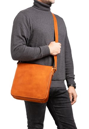 Man draagt een grijze coltrui en zwarte broek en houdt een oranje leren schoudertas met een riem over de borst vast.