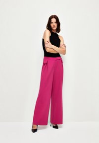 Pantalons large fuchsia taille haute avec poches avant et un détail en nœud ; associés avec un top sans manches noir et des talons noirs.