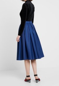 Sininen laskostettu hame, joka on valmistettu sileästä kankaasta, ja jossa on korkea vyötärö ja liehuva siluetti. Yhdistetty istuvaan mustaan samettitoppaukseen ja korkokenkiin.