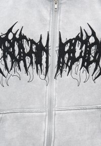 Γκρι φούτερ με φερμουάρ και μεγάλο μαύρο κεντημένο σχέδιο με αφηρημένες, αιχμηρές μορφές στο στήθος. Μαλακή υφή υφάσματος.