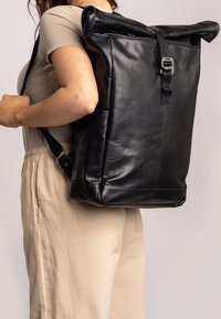 Čierny kožený ruksak so skladacím vrchom, nastaviteľnými popruhmi a kovovou prackou, s hladkou textúrou a minimalistickým dizajnom.