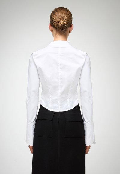 Blusa bianca aderente con maniche lunghe, orlo leggermente curvato e cuciture posteriori. Abbinata a una gonna nera a vita alta con due tasche posteriori.