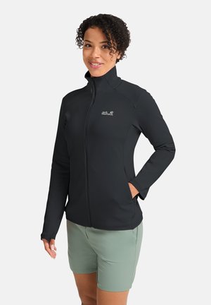 Frau trägt eine schwarze Jacke mit Reißverschluss und hohem Kragen sowie grüne Shorts, steht mit einer Hand in der Tasche vor einem schlichten Hintergrund.